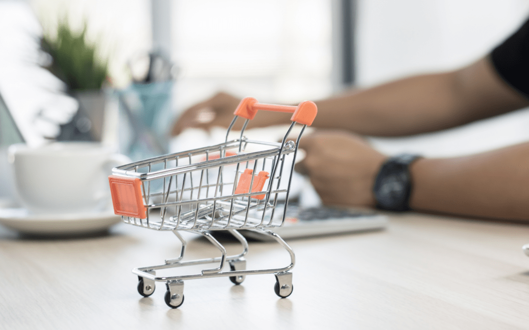 Ein kleiner Einkaufswagen steht auf einem Tisch neben einer Kaffeetasse und einem Laptop. Der Einkaufswagen symbolisiert das Thema E-Commerce.