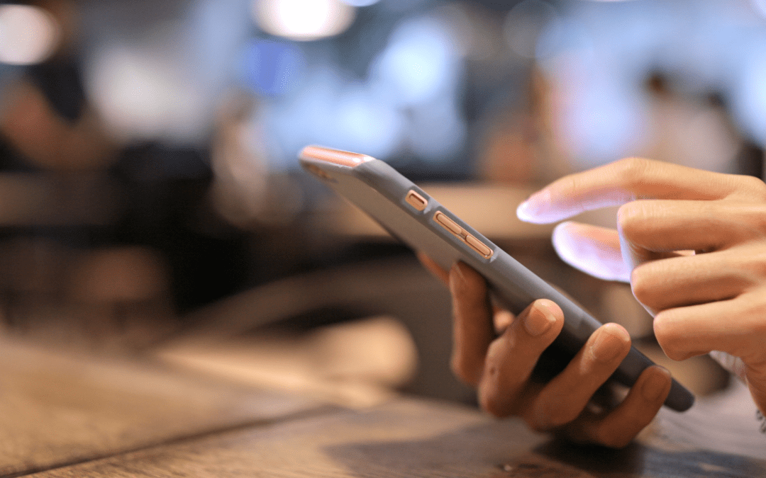 Eine Person nutzt ein Smartphone in einem gemütlichen Café; der Daumen tippt auf den Bildschirm, während der andere Hand eine leichte Unterstützung gibt.