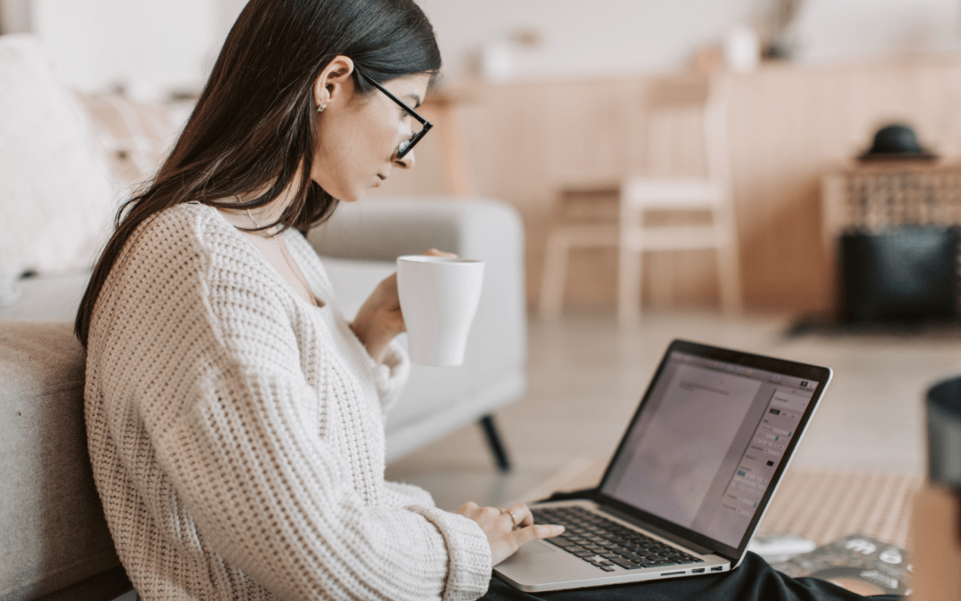 Eine fokussierte Person sitzt auf dem Boden, arbeitet am Laptop und hält eine Tasse in der Hand. Der Raum ist modern und freundlich gestaltet.