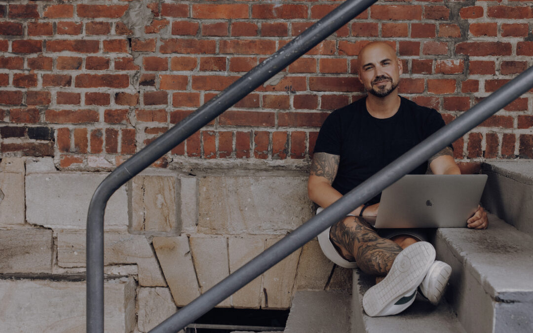 Ein Mann sitzt entspannt auf einer Treppe mit einem Laptop. Sein Outfit und Tatoos verleihen ihm einen lässigen Look in einem urbanen Ambiente.